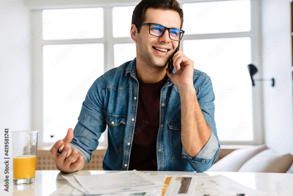 Joyful brunette man talking on cellphone while working with papers