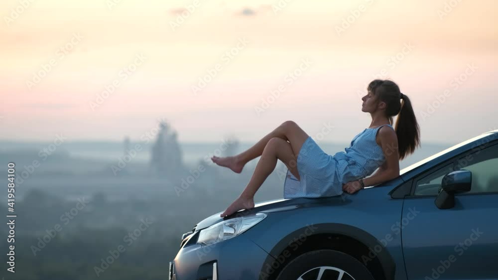 Happy young woman in blue dress sitting on her vehicle bonnet looking at sunset view of summer nature. Travelling and vacation concept. Happy young woman in blue dress sitting on her vehicle bonnet looking at sunset view of summer nature. Travelling and vacation concept.