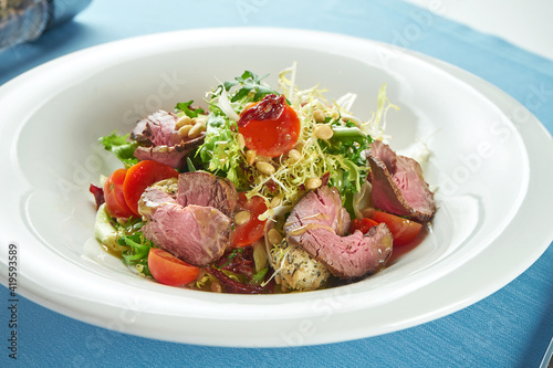 Appetizing, warm salad with roast beef, cherry tomatoes, parmesan and pine nuts in a white plate on a blue tablecloth