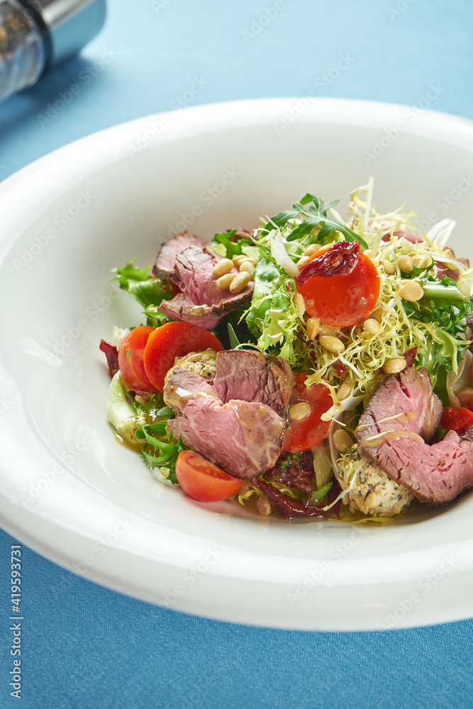 Appetizing, warm salad with roast beef, cherry tomatoes, parmesan and pine nuts in a white plate on a blue tablecloth Appetizing, warm salad with roast beef, cherry tomatoes, parmesan and pine nuts in a white plate on a blue tablecloth