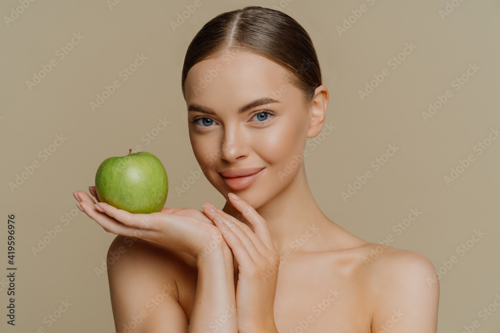 Photo of shirtless brunette young woman with combed hair holds fresh green apple uses natural cosmetic has bare shoulders isolated over brown background. People cosmetology body care concept Photo of shirtless brunette young woman with combed hair holds fresh green apple uses natural cosmetic has bare shoulders isolated over brown background. People cosmetology body care concept