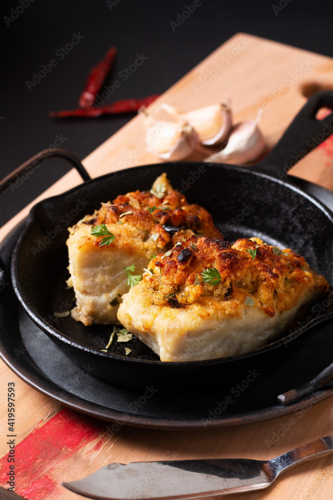 Food concept spot focus homemade butter, garlic, bread crumbs and cheese crunchy Baked cod fish in skillet iron cast pan on black background with copy space