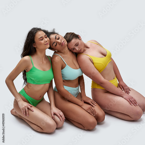 Three diverse young women in colorful underwear leaning on each other, sitting on knees with eyes closed isolated over white background