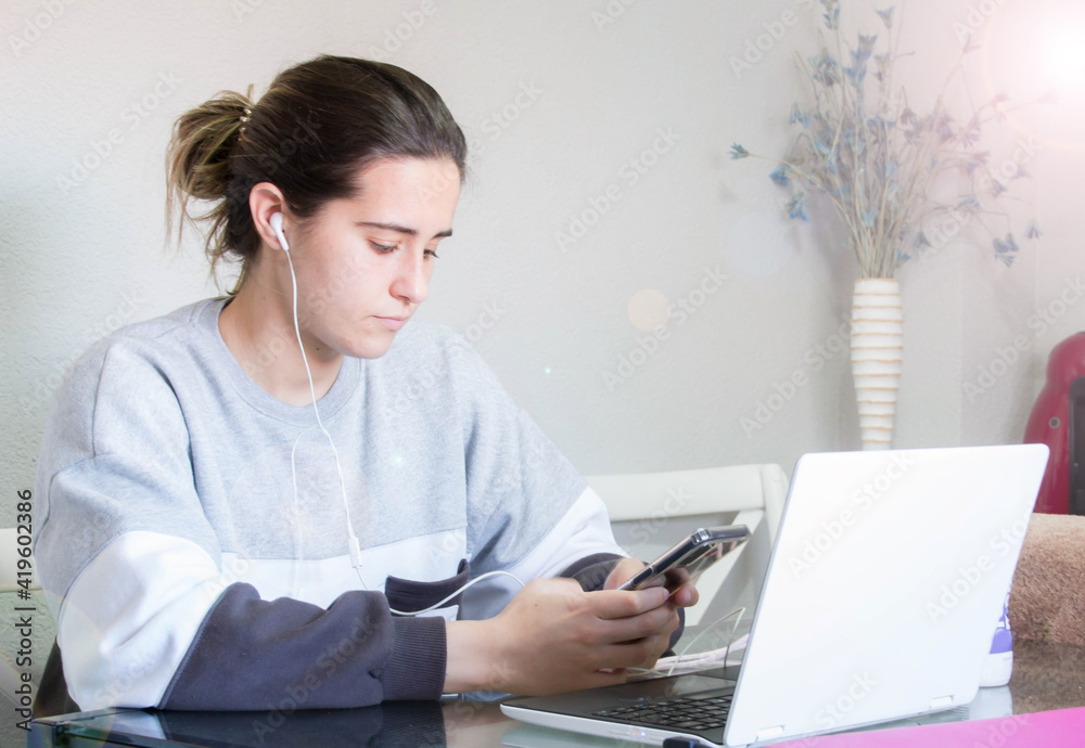 Attractive cheerful young brunette businesswoman sitting at the table with laptop computer indoors, wearing wireless earphones, audio call, waving hand Attractive cheerful young brunette businesswoman sitting at the table with laptop computer indoors, wearing wireless earphones, audio call, waving hand