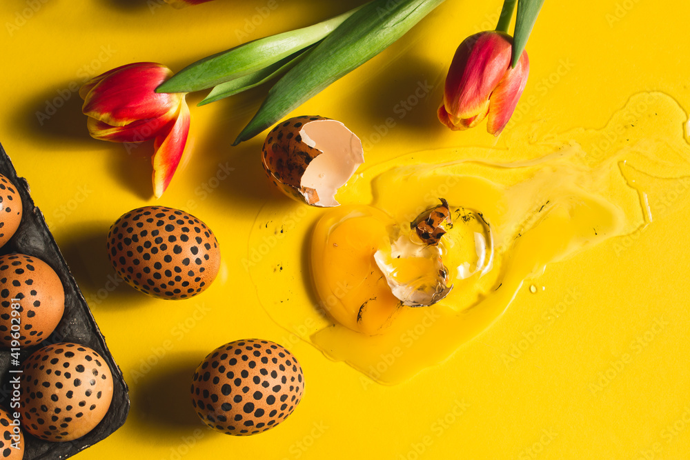 Broken painted easter egg on the yellow surface. Painted easter eggs with black dots in an egg box with tulips. Close up easter flat lay. Trend color of the year 2021 Broken painted easter egg on the yellow surface. Painted easter eggs with black dots in an egg box with tulips. Close up easter flat lay. Trend color of the year 2021
