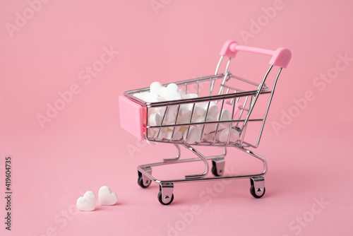 A supermarket cart filled to the brim with white candy hearts on a pink background. The concept of buying presents.