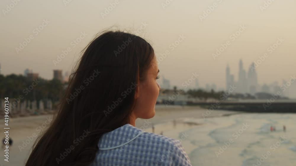 Woman looking at sand beach and city skyline across the water. Back view of attractive happy woman with long brown hair looking at Dubai skyline during sunset Woman looking at sand beach and city skyline across the water. Back view of attractive happy woman with long brown hair looking at Dubai skyline during sunset