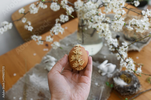 Hand holding Easter egg decorated with dry flower at rustic table with linen napkin, cherry blossom