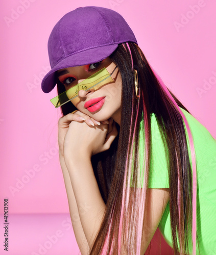 Obraz na płótnie Portrait of young brunette woman with colored hair strands wearing green shirt, cap and stylish rectangular sunglasses holding chin in hands over pink background. Fashion, style, hair coloring concept w salonie