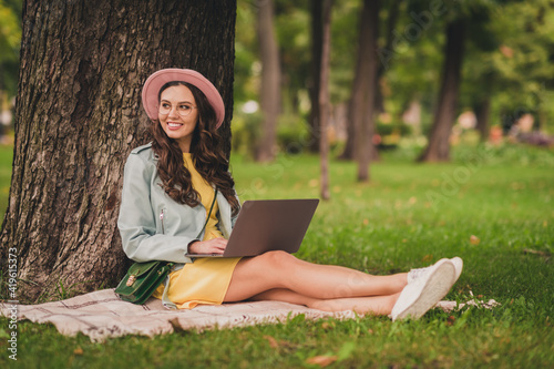 Photo of charming young woman dressed teal jacket glasses bag cap sitting green grass writing modern device outdoors city park