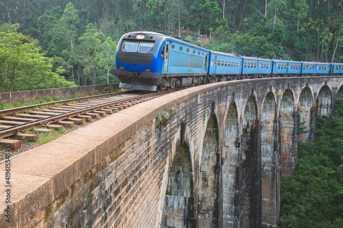 A blue train crosses the Demodara Nine Arches Bridge viaduct passing through the jungle hill country of Sri Lanka on a scenic journey.