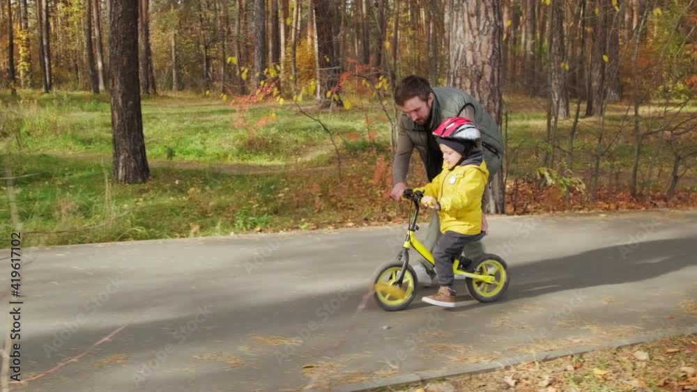 Tracking shot of father teaching cute 5-year-old son how to ride balance bicycle in park Tracking shot of father teaching cute 5-year-old son how to ride balance bicycle in park