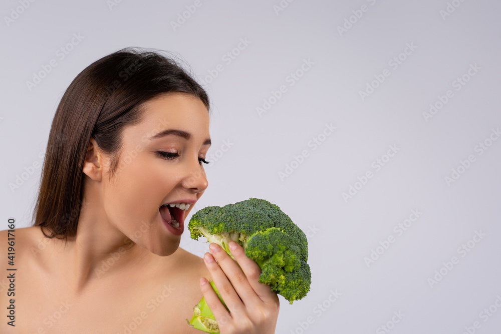 Portrait of a delighted young , naked woman eating broccoli against a gray background. Side space for advertisement Portrait of a delighted young , naked woman eating broccoli against a gray background. Side space for advertisement
