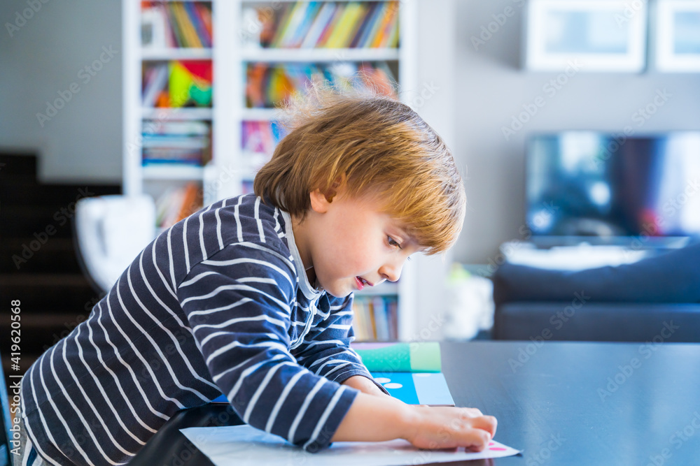 Little boy wearing striped t shirt sitting at the table, learning at home during coronavirus quarantine. Toddler glueing stickers in the early development book. Child educating in playing. Little boy wearing striped t shirt sitting at the table, learning at home during coronavirus quarantine. Toddler glueing stickers in the early development book. Child educating in playing.