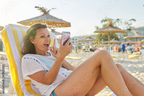 Happy middle-aged woman talking on smartphone using video call, on beach