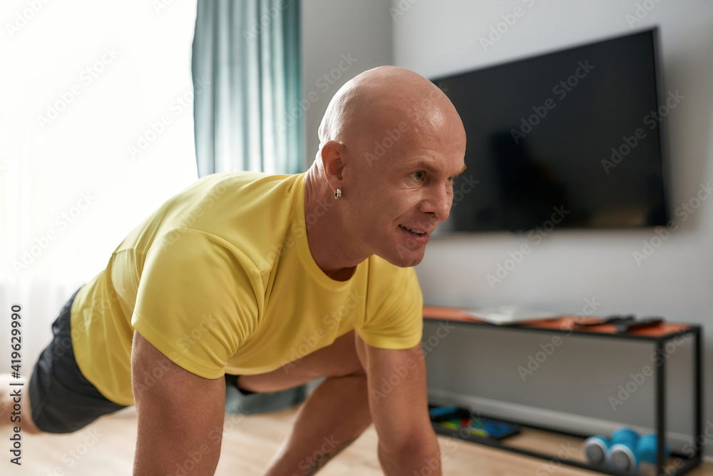 Fototapeta premium Portrait of bald man, fitness coach stretching his body, working out in living room at home on a daytime