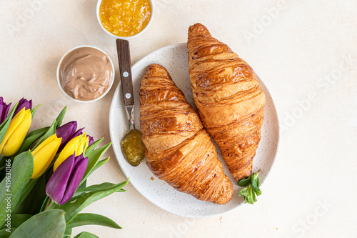 Breakfast with fresh croissants, chocolate cream, jam and colorful tulips on light stone background. Beautiful romantic composition. Top view.