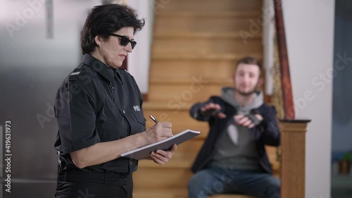 Side view of serious Caucasian policewoman in sunglasses filling in documents as caught robber talking and gesturing at background. Police officer arresting thief at crime scene.