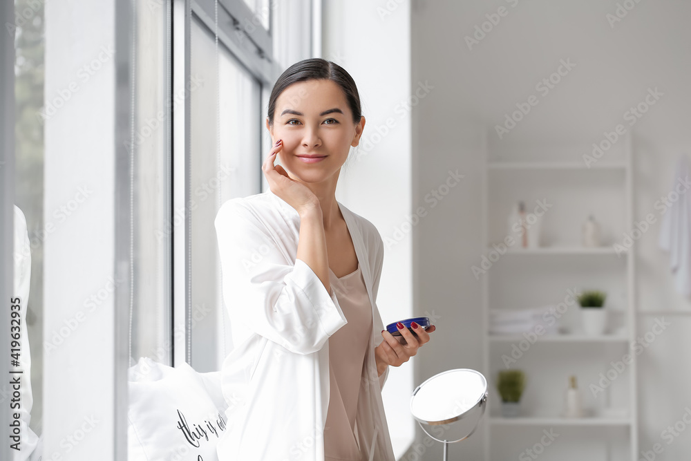 Beautiful young woman applying facial cream in bathroom