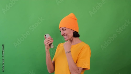 Hipster woman in orange hat green background amazed by unbelievable shopping mobile app sale message looking at smartphone, girl winner holding cell phone screaming with joy 