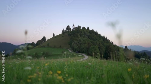 Slow motion shot of a grass field and flowers in front of a hill with a church on top. Shot in the morning at blue hour just before sunrise at Sveti Jakob, Medvode, Slovenia. photo