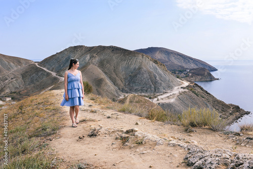 Girl with a blue dress walks along a mountain path and looks at the sea. Walking in the highlands. High mountains and sea views. Summer rest. Tourism and travel