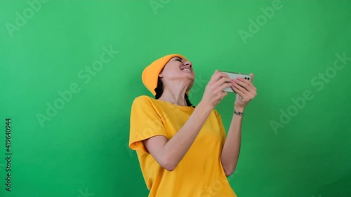 Hipster woman in orange hat green background smiling plays games on mobile phone, holding phone horizontal winner emotions 