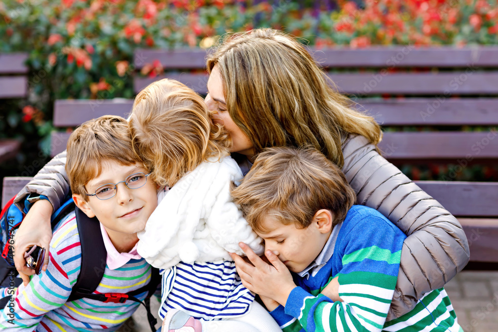 Fototapeta premium Mother and three children hugging. Happy family sitting outdoor: woman and two brother kids boys and cute little toddler girl. Siblings and mum in love. Healthy people giving hugs.