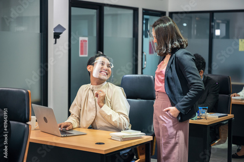 Asian businesswoman consulting with happy and smiling colleague wearing face shield at desk in modern office