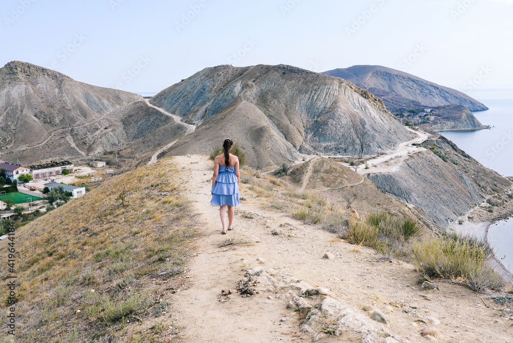 Walking in the highlands. A girl with a blue dress walks along a mountain path overlooking the sea. Tourism, travel and outdoor activities