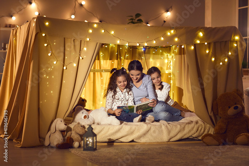 Happy family enjoying a good quiet evening at home. Mommy telling fairy tales to children. Young mother and two little daughters reading book of bedtime stories sitting in cozy playroom tent together