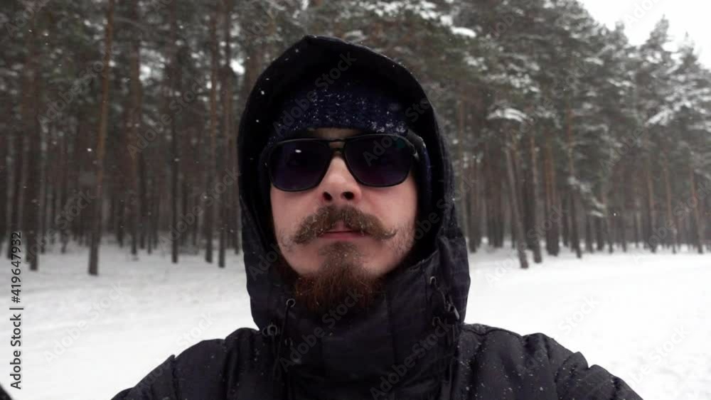 A young bearded man in sun glasses is making selfie standing in a pine wood among falling snow in slow motion. Portrait of a serious man walking in winter forest. Theme of spending free time outdoors. A young bearded man in sun glasses is making selfie standing in a pine wood among falling snow in slow motion. Portrait of a serious man walking in winter forest. Theme of spending free time outdoors.