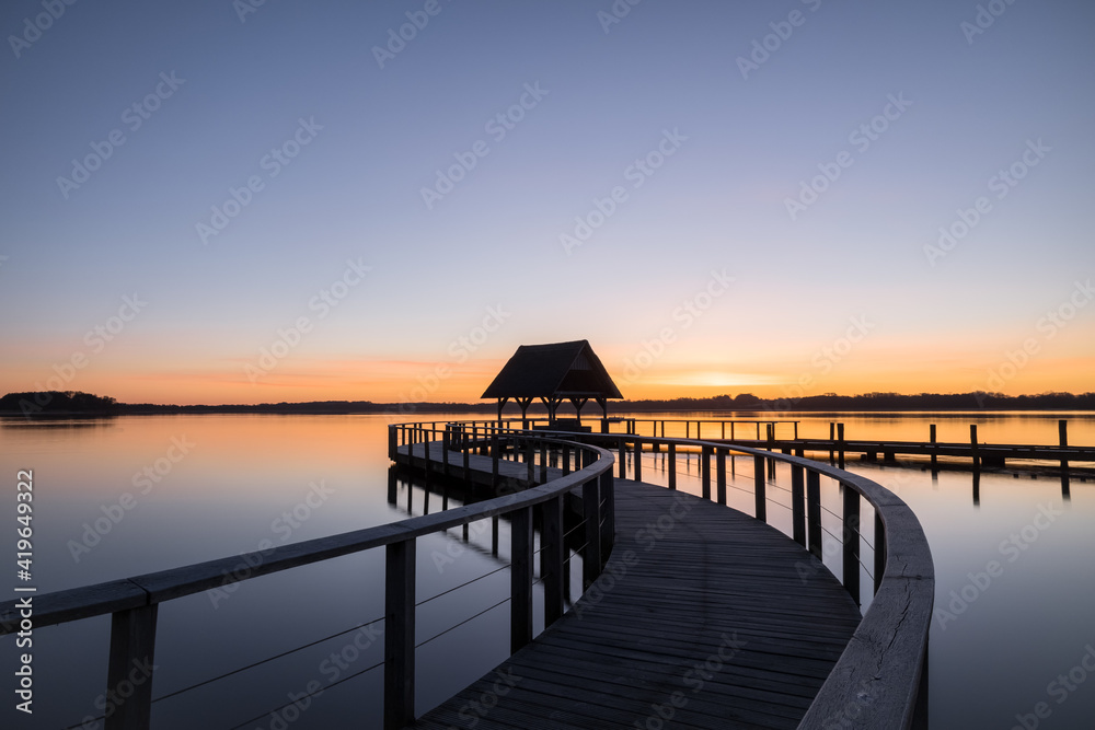 Swinged Pier construction and shelter with thatched roof at beautiful colorful sunrise under clear sky at Lake Hemmelsdorf, Schleswig-Holstein, Northern Germany