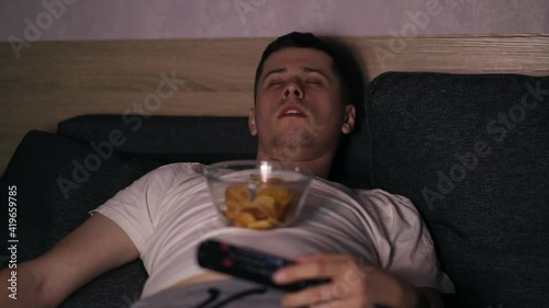A man lying at coach and watching TV, eating fastfood