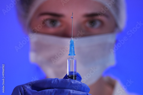 Doctor in sterile cap, protective mask and blue medical gloves preparing syringe for injection, selective focus