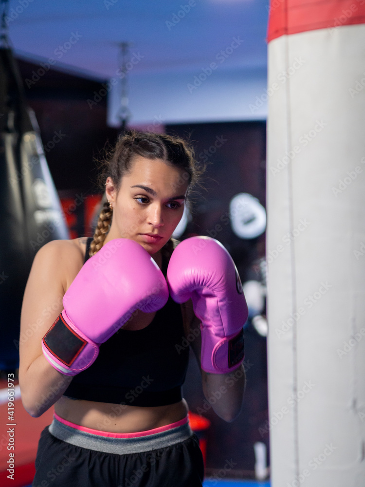 Woman boxing with pink gloves and focused on a punching bag in a gym. Young female with french braids training with confidence for protection. Cancer fight and girl power concepts.