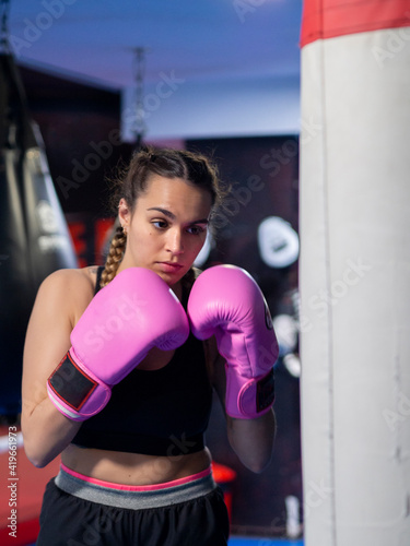 Woman boxing with pink gloves and focused on a punching bag in a gym. Young female with french braids training with confidence for protection. Cancer fight and girl power concepts. © OlgaPS