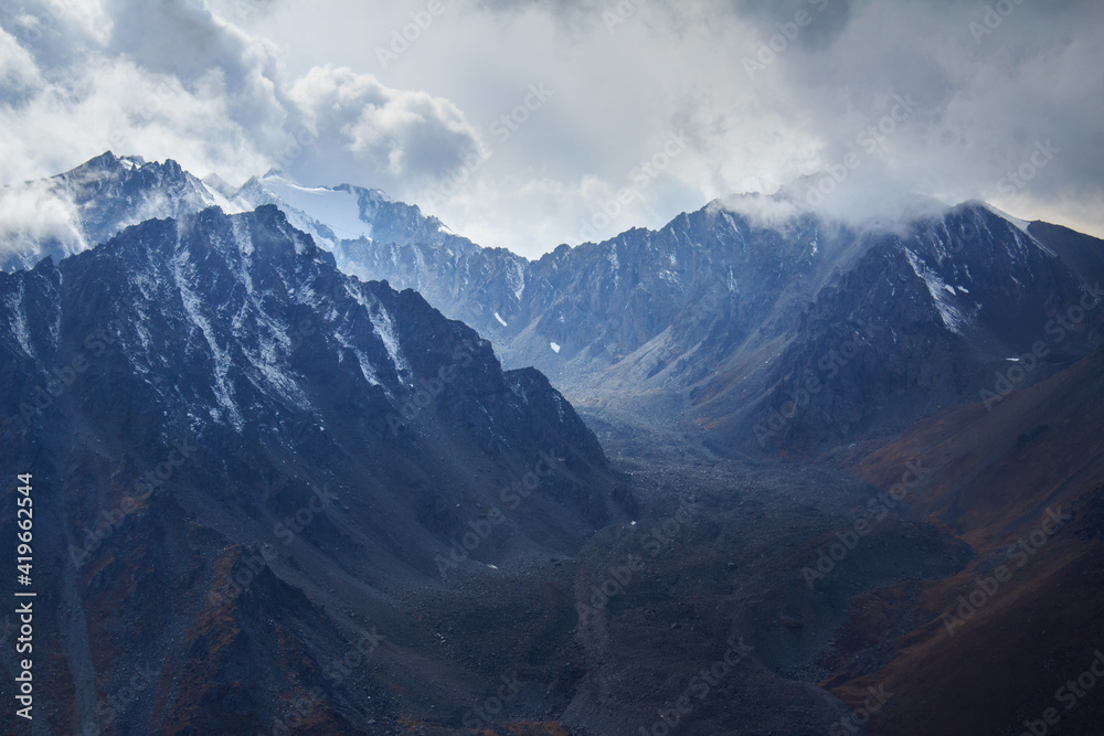 Snow lies on the sharp rocky peaks of the mountain range in the Prohodnoe gorge, beautiful relief mountain slopes, stone talus on the slopes, a sky with thick dark clouds, autumn, cloudy, sunlight
