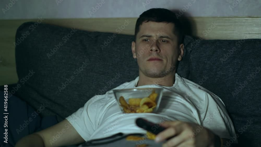 A man lying at coach and watching TV, eating fastfood A man lying at coach and watching TV, eating fastfood