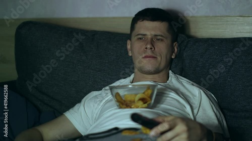 A man lying at coach and watching TV, eating fastfood