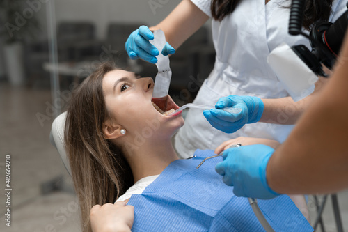 A young woman of Caucasian ethnicity sits in a chair in a dental clinic with an open mouth. The hands of the nurse and the orthodontist manipulate the patient's mouth.