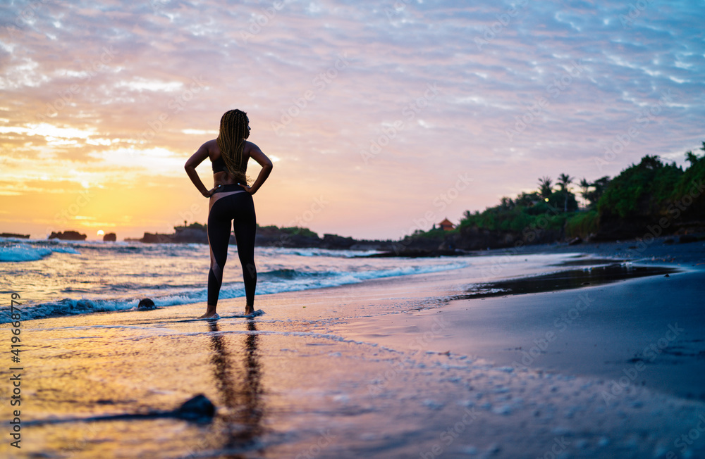 Back view of fit girl in tracksuit looking at scenery landscape spending evening time for enjoying breathtaking sunset and running across seashore coastline, woman with casual figure near ocean