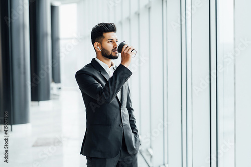 Modern Indian man in a black suit drinks his coffee in the middle of a modern building, office worker, boss, businessman