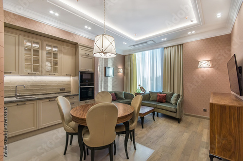 Spacious kitchen with the large lounge area with a soft velvet olive sofa. Neoclassic beige kitchen with built-in appliances. The round table with soft chairs standing in the center of the kitchen