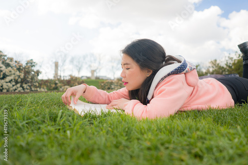 young Asian woman enjoying novel on grass - lifestyle portrait of young happy and pretty Japanese girl reading a book at beautiful city park in reading and studying concept