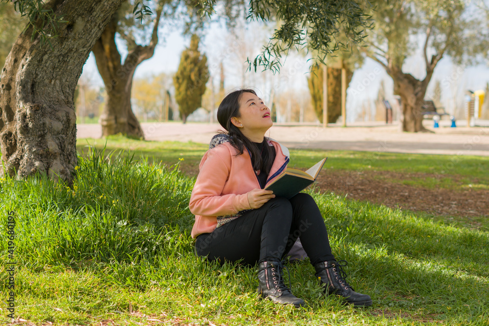 young Asian woman enjoying novel on grass - lifestyle portrait of young happy and pretty Korean girl reading a book at beautiful city park in reading and studying concept