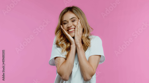 Canvastavla flattered woman touching cheeks and smiling isolated on pink