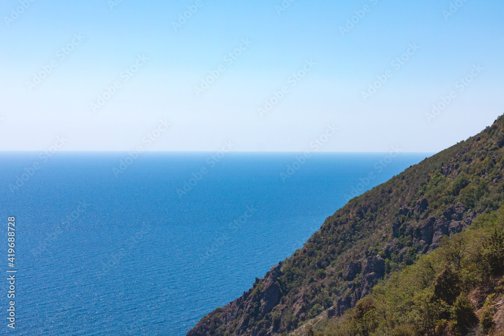 Rocky mountain slope covered with green plants against the backdrop of the blue sea and cloudless blue gradient sky