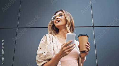 Foto low angle view of blonde woman holding coffee to go and smartphone near building
