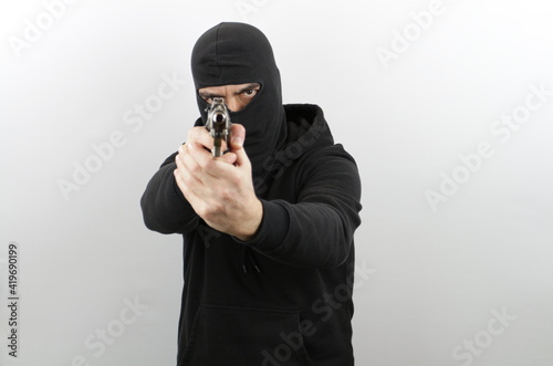 Man in black mask holding pistol and pointing at the cameraover white background. Crime concept